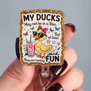 May include: A glittery badge reel with the text "MY DUCKS May not be in a Row, But at least They are having FUN." The badge features a cartoon duck wearing sunglasses and a bow, holding a drink. Butterflies and flowers surround the duck.