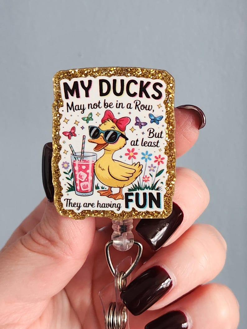 May include: A glittery badge reel with the text "MY DUCKS May not be in a Row, But at least They are having FUN." The badge features a cartoon duck wearing sunglasses and a bow, holding a drink. Butterflies and flowers surround the duck.