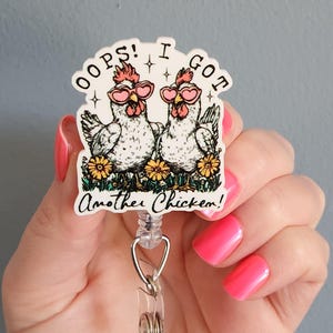 May include: A white and black badge reel featuring two chickens wearing heart-shaped sunglasses, with the text "Oops! I got Another Chicken!" in a whimsical font. The chickens are surrounded by yellow flowers.
