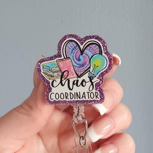 May include: A badge reel featuring a purple glitter border and a central graphic with a tie-dye heart, a stack of books, and a lightbulb. The text reads chaos COORDINATOR in black script and block lettering on a white background.