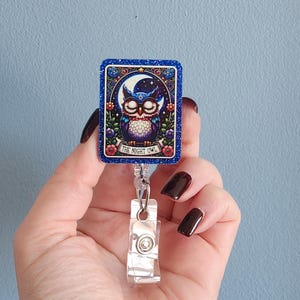 May include: A rectangular badge reel with a glittery blue border and a detailed illustration of an owl under a crescent moon. The text "THE NIGHT OWL" is at the bottom. The reel has a clear plastic clip and retractable cord.