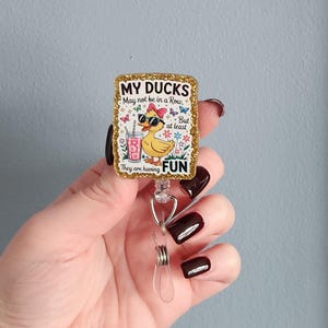 May include: A rectangular badge reel with a gold glitter border. The badge features a cartoon duck wearing sunglasses and a bow, with the text "MY DUCKS May not be in a Row, But at least They are having FUN."