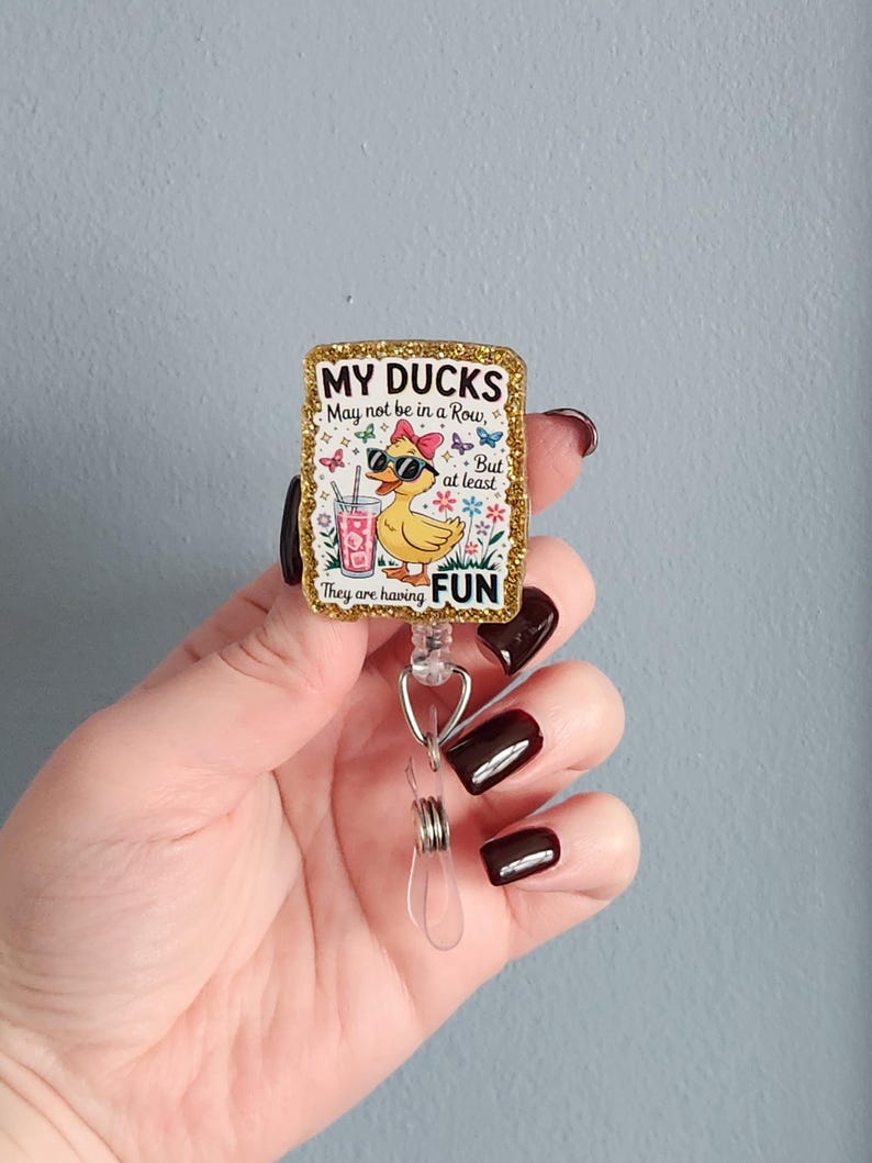 May include: A rectangular badge reel with a gold glitter border. The badge features a cartoon duck wearing sunglasses and a bow, with the text "MY DUCKS May not be in a Row, But at least They are having FUN."