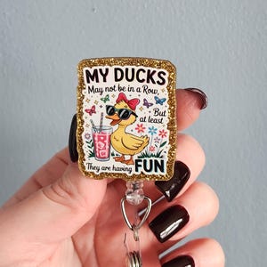 May include: A rectangular badge reel with a glittery gold border. The badge features a cartoon duck wearing sunglasses and a bow, holding a drink. Text reads "MY DUCKS May not be in a Row, But at least They are having FUN."