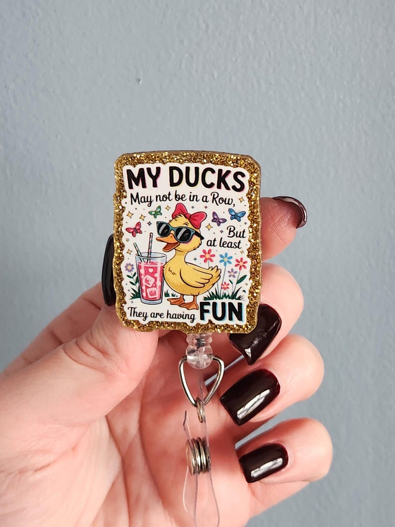 May include: A rectangular badge reel with a glittery gold border. The badge features a cartoon duck wearing sunglasses and a bow, holding a drink. Text reads "MY DUCKS May not be in a Row, But at least They are having FUN."