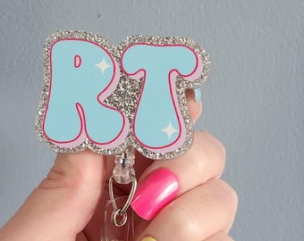 May include: A badge reel featuring a glittery silver border and the letters RT in light blue with a pink outline. The letters are decorated with small white star accents. The badge reel has a clear plastic retractable cord and a metal clip attachment.
