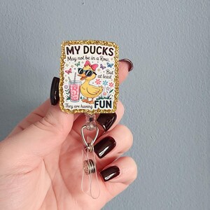 May include: A gold glitter badge reel with the text "MY DUCKS May not be in a Row, But at least They are having FUN." The badge features a cartoon duck wearing sunglasses and a bow, holding a drink.