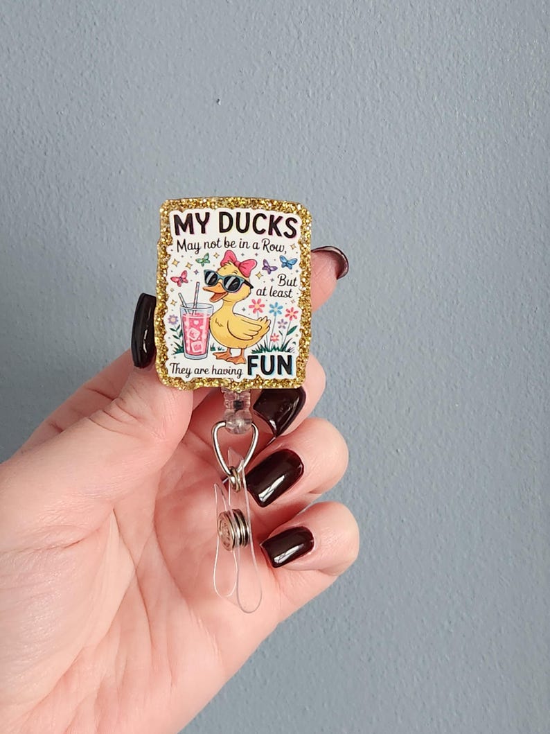 May include: A gold glitter badge reel with the text "MY DUCKS May not be in a Row, But at least They are having FUN." The badge features a cartoon duck wearing sunglasses and a bow, holding a drink.