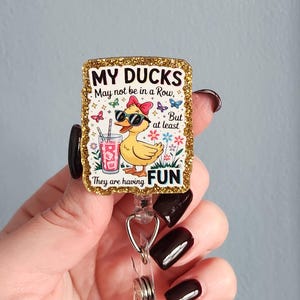 May include: A rectangular badge reel with a gold glitter border. The badge features a cartoon duck wearing sunglasses and a bow, holding a drink. Text reads "MY DUCKS May not be in a Row, But at least They are having FUN."