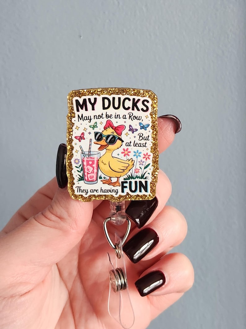 May include: A rectangular badge reel with a gold glitter border. The badge features a cartoon duck wearing sunglasses and a bow, holding a drink. Text reads "MY DUCKS May not be in a Row, But at least They are having FUN."