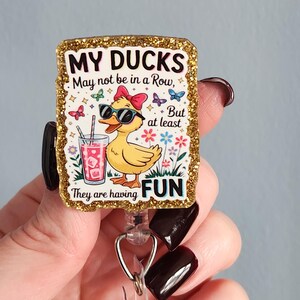 May include: A retractable badge reel with a gold glitter border. The badge reel features a cartoon duck wearing sunglasses and a bow, holding a drink. Text reads "MY DUCKS May not be in a Row, But at least They are having FUN."