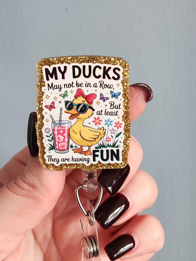 May include: A retractable badge reel with a gold glitter border. The badge reel features a cartoon duck wearing sunglasses and a bow, holding a drink. Text reads "MY DUCKS May not be in a Row, But at least They are having FUN."
