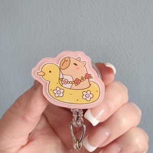 May include: A pink and yellow badge reel featuring a cartoon capybara floating in a yellow duck-shaped floatie. The capybara wears a flower lei. The badge reel has a retractable cord and a metal clip.