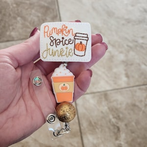 May include: A white glitter badge reel with a white rectangle that says "Pumpkin Spice Junkie" with a brown coffee cup with a pumpkin inside. The coffee cup is attached to a gold bead and a clear retractable badge holder.