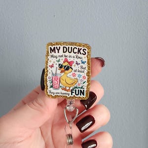 May include: A rectangular badge reel with a gold glitter border. The badge features a cartoon duck wearing sunglasses and a pink bow, with the text "MY DUCKS May not be in a Row, But at least They are having FUN."