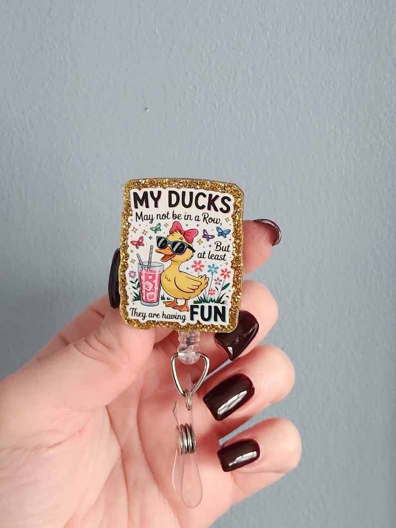 May include: A rectangular badge reel with a gold glitter border. The badge features a cartoon duck wearing sunglasses and a pink bow, with the text "MY DUCKS May not be in a Row, But at least They are having FUN."