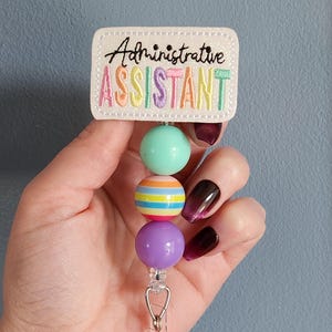 May include: A retractable badge reel with the text "Administrative ASSISTANT" embroidered in colorful thread on a white rectangular patch. The reel features three colorful beads: mint green, striped, and purple.