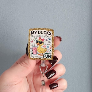 May include: A retractable badge reel with a rectangular design. The design features a cartoon duck wearing sunglasses and a bow, holding a drink. The text reads "MY DUCKS May not be in a Row, But at least They are having FUN". The border is gold glitter.