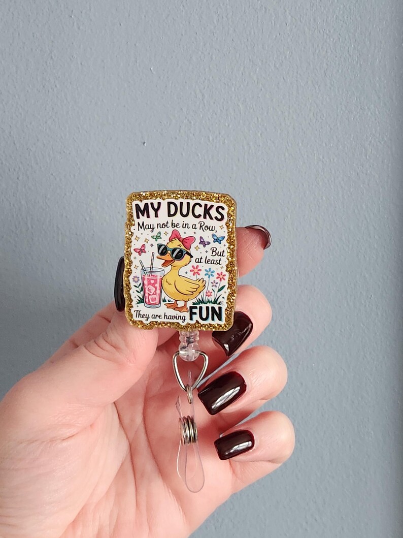 May include: A retractable badge reel with a rectangular design. The design features a cartoon duck wearing sunglasses and a bow, holding a drink. The text reads "MY DUCKS May not be in a Row, But at least They are having FUN". The border is gold glitter.