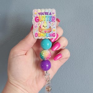 May include: A badge reel featuring a rectangular acrylic topper with the text YOU'RE A GLITTER DUMPLING and a cartoon dumpling illustration. Below the topper are three beads: one solid teal, one multicolored glitter, and one solid purple bead.