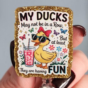 May include: A rectangular badge with a gold glitter border. The badge features a yellow duck wearing sunglasses and a red bow, holding a drink. Text reads "MY DUCKS May not be in a Row, But at least They are having FUN."