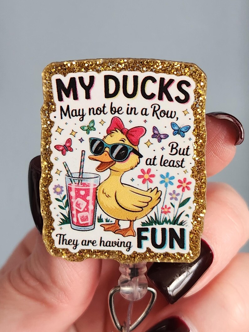 May include: A rectangular badge with a gold glitter border. The badge features a yellow duck wearing sunglasses and a red bow, holding a drink. Text reads "MY DUCKS May not be in a Row, But at least They are having FUN."