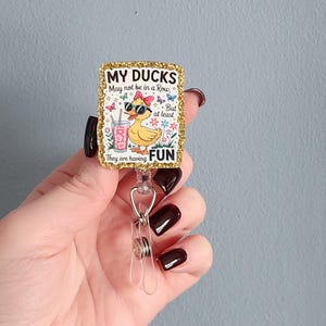 May include: A rectangular badge reel with a gold glitter border. The badge features a cartoon duck wearing sunglasses and a bow, holding a drink. Text reads "MY DUCKS May not be in a Row, But at least They are having FUN."