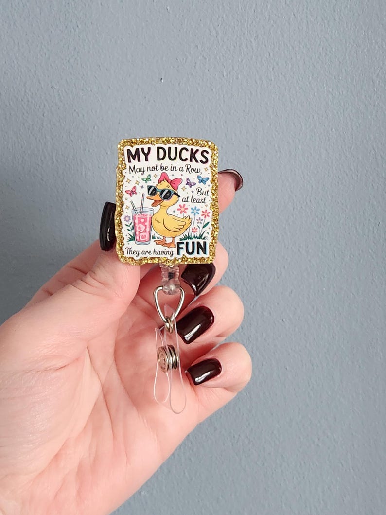 May include: A rectangular badge reel with a gold glitter border. The badge features a cartoon duck wearing sunglasses and a bow, holding a drink. Text reads "MY DUCKS May not be in a Row, But at least They are having FUN."