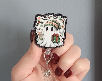 May include: A white ghost-shaped badge reel with a Christmas theme. The ghost wears a green and red hat and holds a gift and a coffee cup. The badge reel has a black glitter border and a clear plastic clip.
