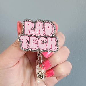 May include: A badge reel featuring a glittery silver border and a pink, bubble-lettered graphic that reads RAD TECH. The design includes small white star accents on the letters and is attached to a clear plastic clip with a metal button mechanism.