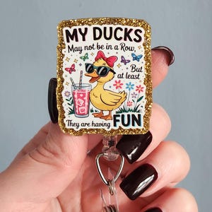May include: A rectangular badge with a gold glitter border features a cartoon duck wearing sunglasses and a bow. The text reads "MY DUCKS May not be in a Row, But at least They are having FUN." The duck is holding a drink with a straw.