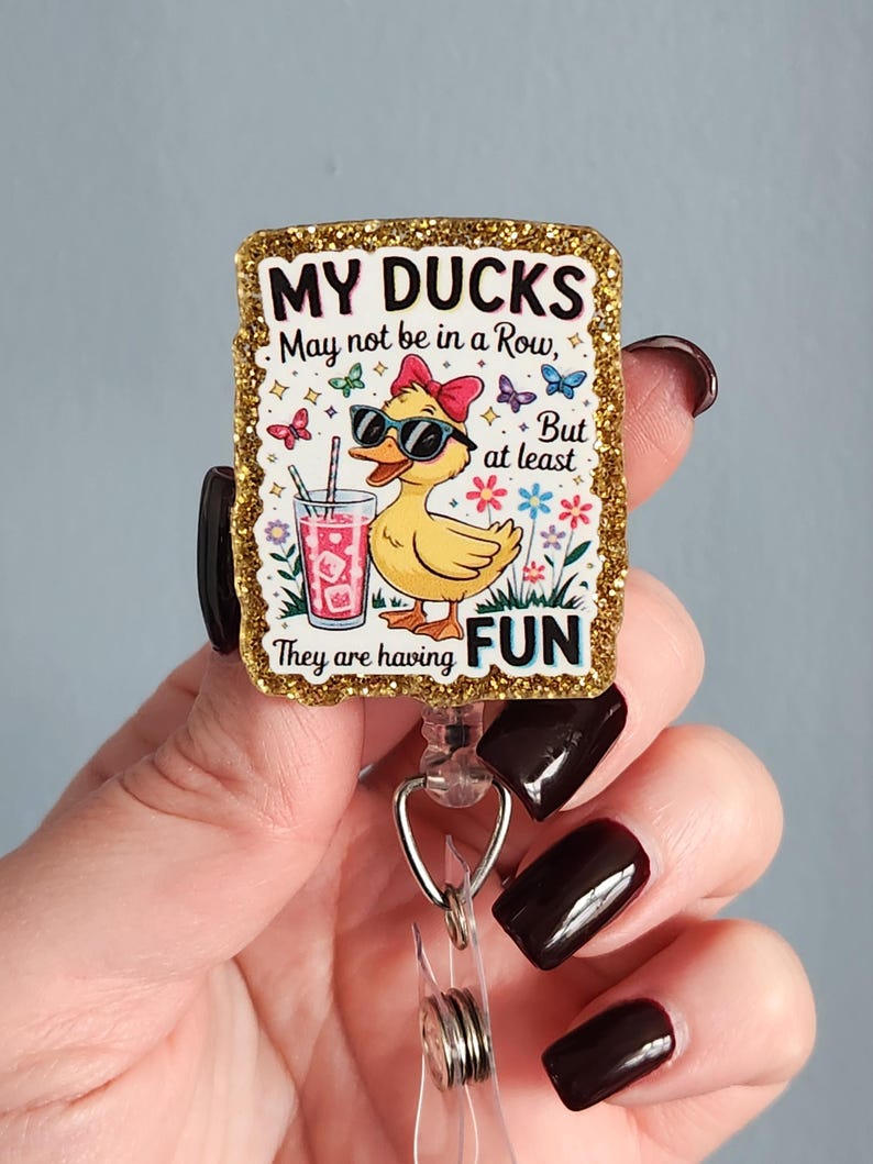 May include: A rectangular badge with a gold glitter border features a cartoon duck wearing sunglasses and a bow. The text reads "MY DUCKS May not be in a Row, But at least They are having FUN." The duck is holding a drink with a straw.