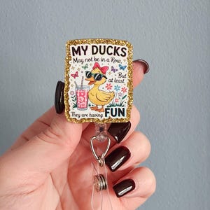 May include: A rectangular badge reel with a gold glitter border. The badge features a cartoon duck wearing sunglasses and a bow, with the text "MY DUCKS May not be in a Row, But at least They are having FUN."