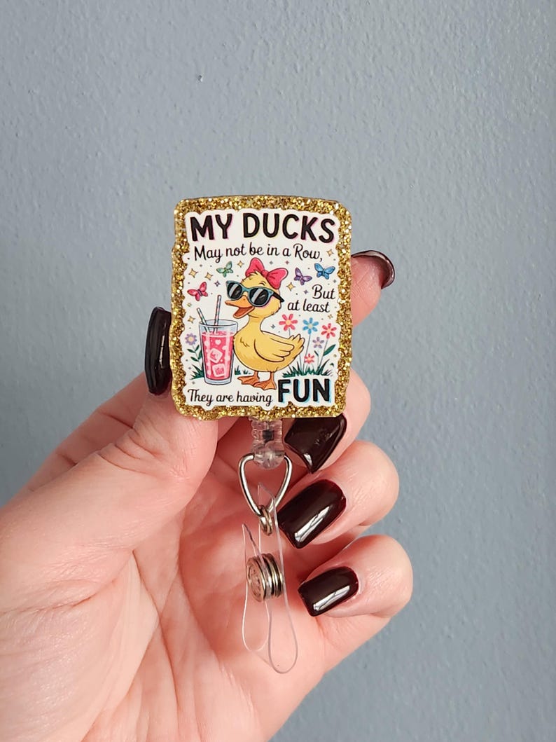 May include: A rectangular badge reel with a gold glitter border. The badge features a cartoon duck wearing sunglasses and a bow, with the text "MY DUCKS May not be in a Row, But at least They are having FUN."