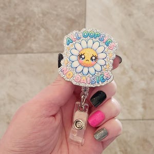 May include: A silver glitter badge holder with a cartoon daisy with a sad face. The daisy has a rainbow colored border and the text "All oopsies no daisies" around it.