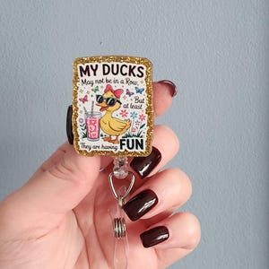 May include: A rectangular badge reel with a gold glitter border. The badge features a cartoon duck wearing sunglasses and a pink bow, holding a drink. Text reads "MY DUCKS May not be in a Row, But at least They are having FUN."