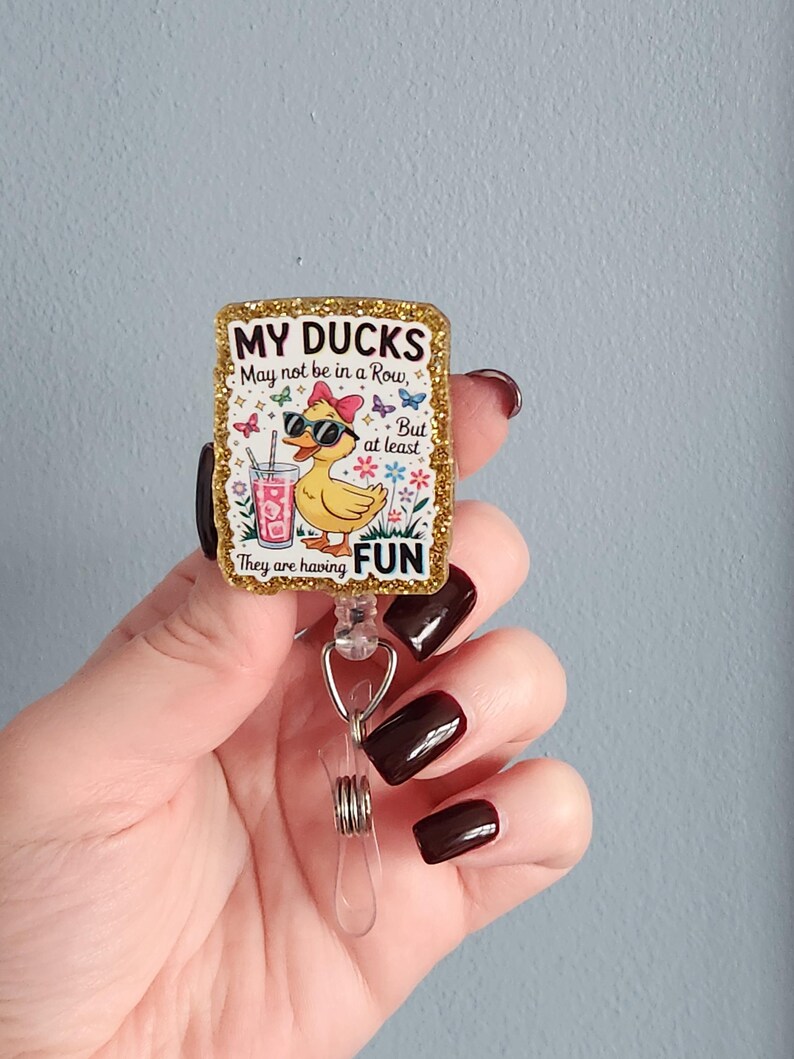 May include: A rectangular badge reel with a gold glitter border. The badge features a cartoon duck wearing sunglasses and a pink bow, holding a drink. Text reads "MY DUCKS May not be in a Row, But at least They are having FUN."