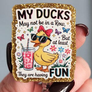 May include: A badge reel with a glittery gold border features a cartoon duck wearing sunglasses and a bow, holding a drink. The text reads "MY DUCKS May not be in a Row, But at least They are having FUN."