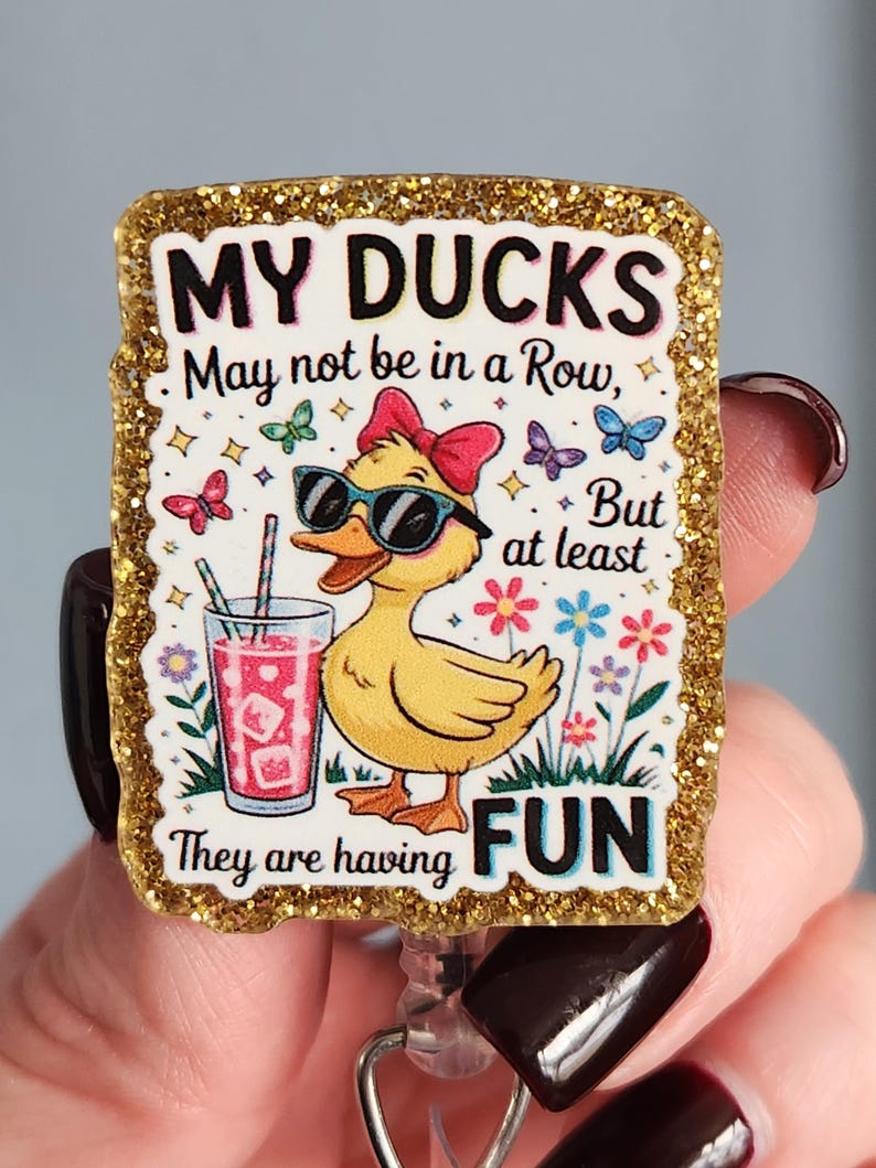 May include: A badge reel with a glittery gold border features a cartoon duck wearing sunglasses and a bow, holding a drink. The text reads "MY DUCKS May not be in a Row, But at least They are having FUN."