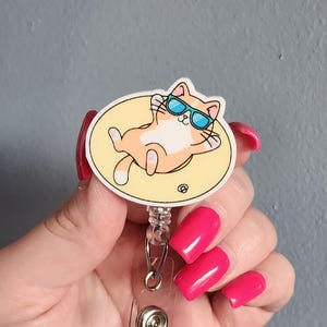 May include: A retractable badge reel featuring a circular graphic of an orange and white cat wearing blue sunglasses while lounging on a yellow circular float. The badge reel has a clear plastic housing with a metal spring mechanism and a metal clip.