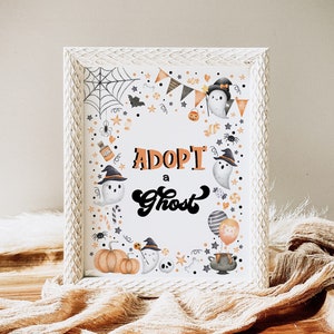 Könnte beinhalten: Weißer Rahmen mit einem weißen Passepartout und einem Druck, der "Adoptiere einen Geist" mit einem Halloween-Thema sagt. Der Druck zeigt Geister, Kürbisse, Zuckerstangen, Fledermäuse, Sterne und ein Spinnennetz.