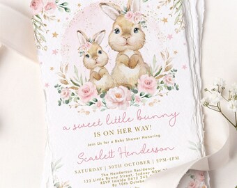 Puede incluir: Una invitación de baby shower rosa y dorada con una ilustración de acuarela de dos conejos con coronas de flores. El texto dice "a sweet little bunny is on her way!" e incluye el nombre "Scarlett Henderson".