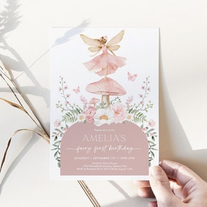 Pode incluir: Um convite de festa de aniversário de fada rosa e branco com uma ilustração em aquarela de uma fada sentada em um cogumelo. O convite inclui o texto "Amelia's fairy first birthday" e detalhes sobre a festa, incluindo a data, a hora e o local.