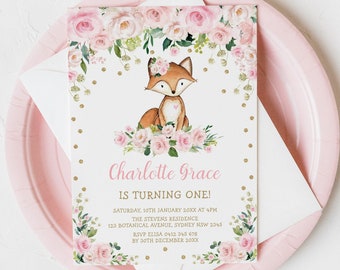 May include: A pink paper plate holds a white envelope and a party invitation. The invitation features a watercolor illustration of a fox with a floral crown and a heart. Text reads "Charlotte Grace is turning one!" with event details.