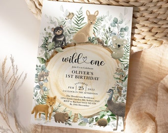Puede incluir: Una ilustración de acuarela de una invitación a una fiesta de animales del bosque para un primer cumpleaños. La invitación presenta un tronco de árbol de madera con el texto "wild one" y un corazón. La invitación también presenta una variedad de animales, incluyendo un canguro, un koala, un wombat, un dingo, un equidna y un emú. La invitación está colocada sobre un fondo de hojas verdes y helechos.
