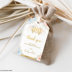 Pu&ograve; includere: Un'etichetta di ringraziamento bianca con lettere dorate e un design ad acquerello di mongolfiere, stelle e fiori. Il testo recita "grazie" e "Per essere venuto e aver festeggiato con noi! Con affetto, Emily e Dan".