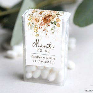 May include: A clear plastic box filled with white mints. The box has a label with a floral design in shades of orange, yellow, and green. The label also includes the text "Mint to Be", "Candace + Alberto", and "15.09.2031".