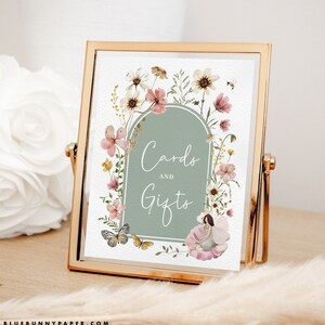 May include: A printable sign with a floral design and a green arch with the text "Cards and Gifts". The sign is in a gold frame and is sitting on a wooden table.