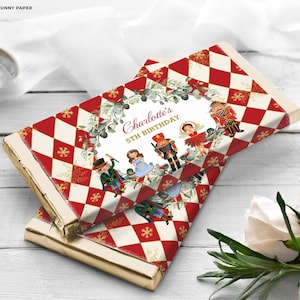 Puede incluir: Envoltorio de barra de chocolate rojo y blanco con borde dorado. El envoltorio presenta un tema de Cascanueces con una bailarina, un soldado Cascanueces y una niña con un vestido azul. El texto en el envoltorio dice "Charlotte's 5th Birthday".