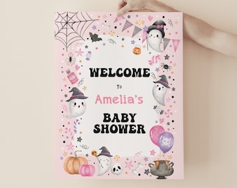 Puede incluir: Un cartel de bienvenida para una baby shower de Halloween rosa y negro con un fantasma, una calabaza y otras decoraciones de Halloween. El cartel dice "Bienvenido a la baby shower de Amelia".